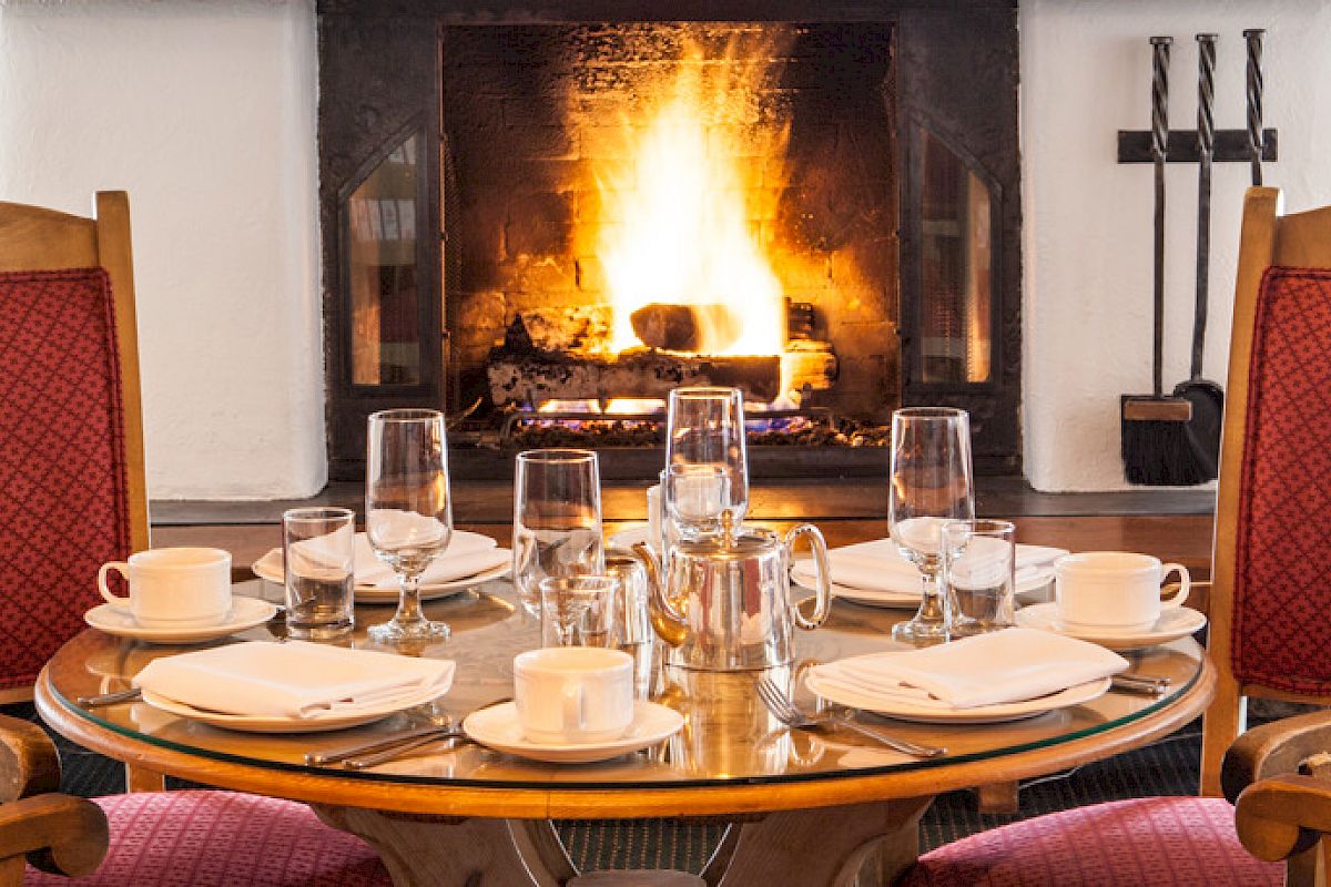 A cozy dining table set for four, located in front of a lit fireplace, with red cushioned chairs and various place settings on the table.