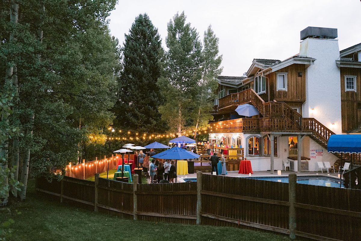 The image shows a lively outdoor gathering with people near a pool, decorative lights, tables, umbrellas, and a large house surrounded by trees and a fence.