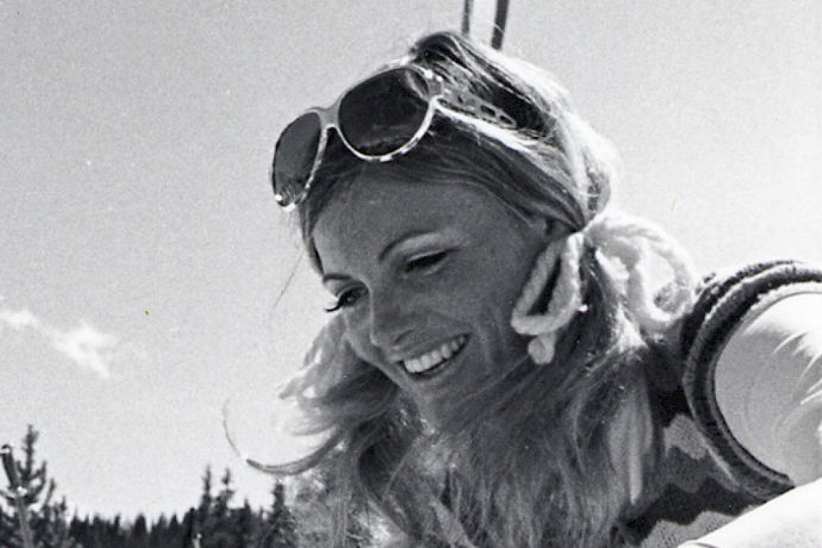 A woman with sunglasses and a smile is in a snowy area with two skis labeled "HEAD Competition" standing upright next to her.