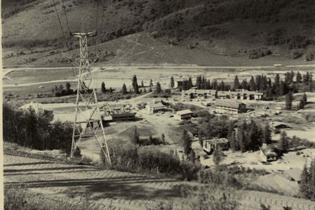 The image shows a small town or settlement in a valley surrounded by hills, with a power line tower in the foreground, and various buildings scattered around.