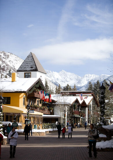 A picturesque village with snow-covered buildings, people walking, and a mountain range in the background under a clear, blue sky.