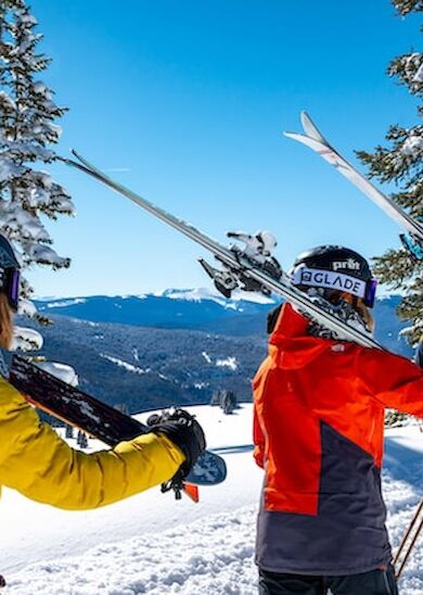 Three people in winter gear carrying skis walk through a snowy landscape surrounded by trees and mountains, under a clear blue sky.