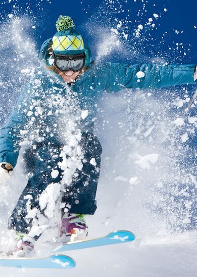 A skier in winter gear is captured mid-motion, amidst a spray of snow, skiing down a snowy slope on a clear day.