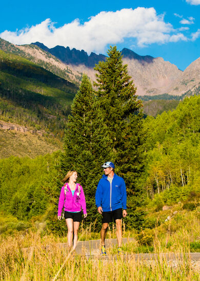 A man and a woman are hiking on a trail surrounded by green trees and mountains under a blue sky with scattered clouds.