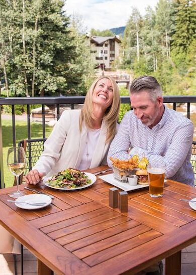 A couple dining outdoors on a patio, with green scenery in the background. A woman smiling and a man looking down at his meal, drinks on the table.