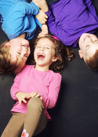 Three children lying on their backs in a circle, laughing and playing together, wearing colorful shirts and enjoying each other's company.