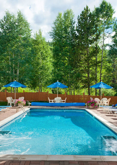 A serene outdoor swimming pool area with lounge chairs and blue umbrellas, surrounded by trees and wooden structures. Clear skies above end the sentence.