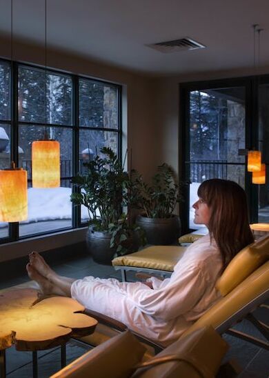 A person in a robe relaxes on a lounge chair in a cozy room with large windows showing a snowy landscape outside and warm hanging lights inside.