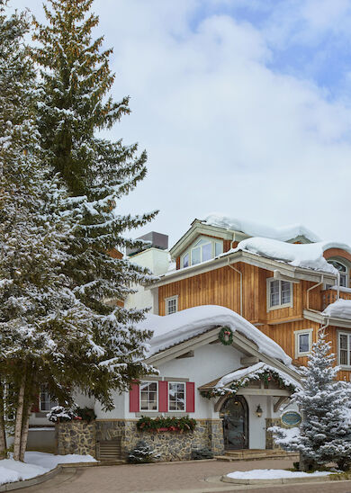 A snowy alpine village scene with wooden chalets, snow-covered trees, and a clear blue sky with clouds, creating a peaceful winter atmosphere.