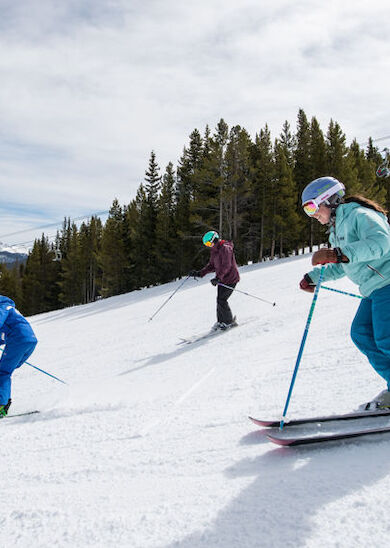 Three people are skiing down a snowy slope with trees and mountains in the background, all wearing helmets and winter gear, enjoying the activity.