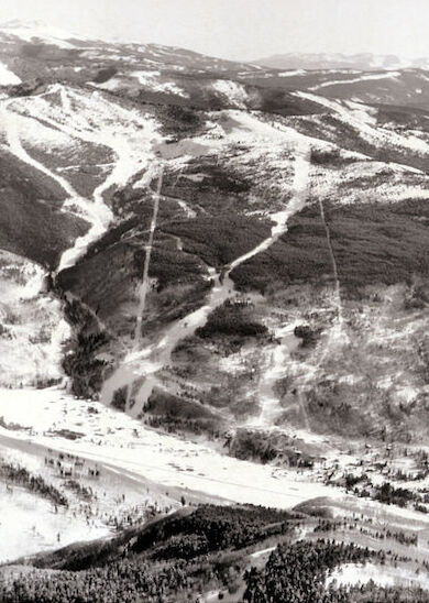 This image shows an aerial view of a snow-covered mountainous area, likely a ski resort, with visible trails and patches of trees.