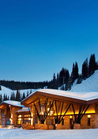 A ski lodge with warm lights illuminates the snowy landscape, set against a backdrop of snow-covered slopes and pine trees under a twilight sky.