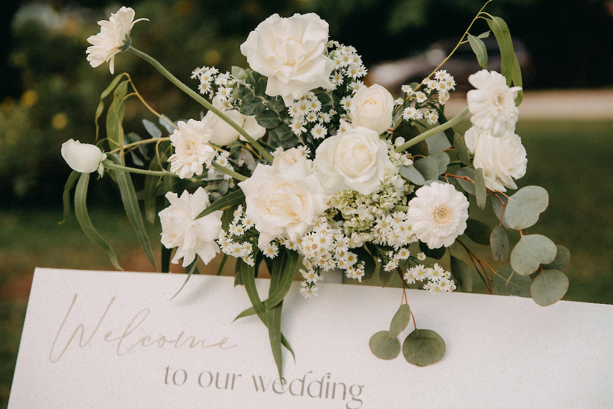 A white floral wedding bouquet with roses and baby's breath sits atop a “Welcome to our wedding” sign, creating a romantic welcome.