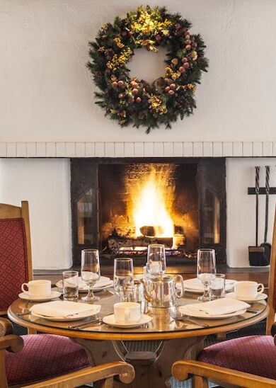 A cozy dining area with a set table for four is in front of a lit fireplace, adorned with a holiday wreath above it and fireplace tools on the side.