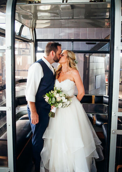 A bride and groom share a kiss inside a modern, glass-enclosed space, with the bride holding a bouquet and the groom in a vest and tie.