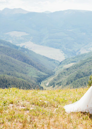 A couple in wedding attire stands on a grassy hilltop, embracing with a scenic mountain valley in the background.