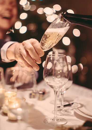 A person is pouring champagne into a glass at a festive dinner table with candles and bokeh lights in the background, while another person laughs.