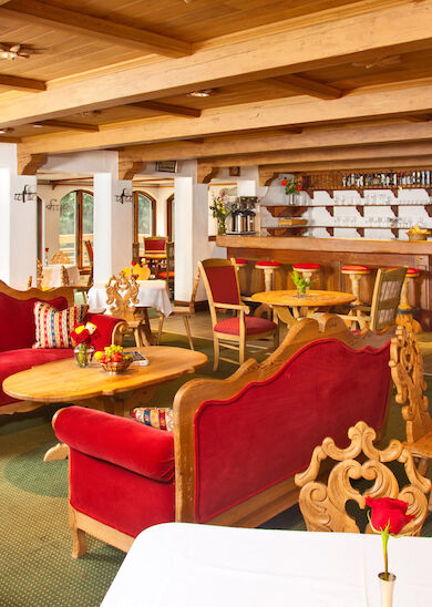 The image shows a cozy, rustic restaurant with red seating, wooden furniture, and tables set with white cloths, decorated with flowers and traditional decor.