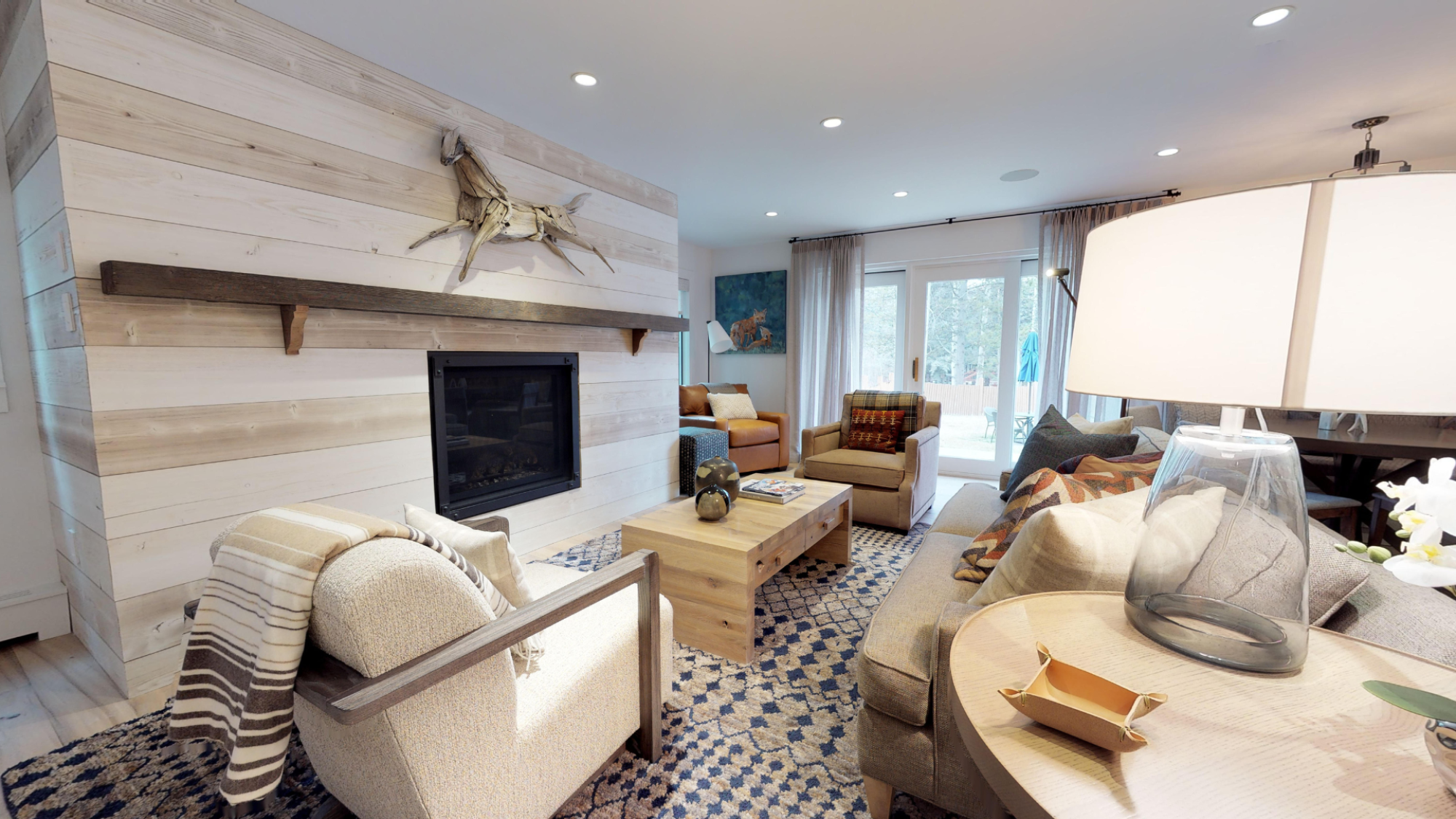 A cozy living room with contemporary furnishings, a fireplace, a wooden coffee table, an armchair, and large windows that let in natural light.