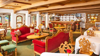 A cozy, rustic restaurant interior with wooden beams, red upholstered seating, ornate wooden chairs, and tables set with white linens and flowers.