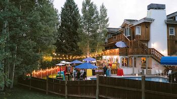 People gathered in a backyard pool area with string lights, umbrellas, and decorations, next to a wooden house surrounded by trees, enjoying an event.