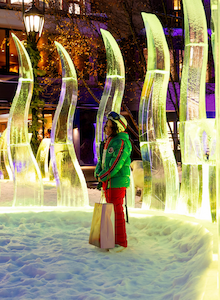 A person stands in a snowy area surrounded by illuminated ice sculptures, holding a paper bag. The scene takes place in a festive, nighttime setting.