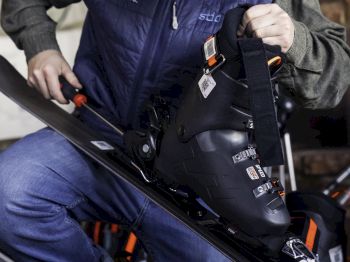 A person is adjusting ski equipment, including ski boots and skis, wearing winter attire and a cap. They appear focused on preparing for skiing.