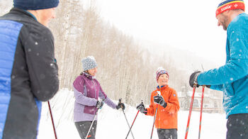 Four people in winter clothing are standing in the snow, holding ski poles and talking.