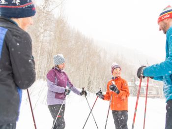 Four people in winter clothing are standing in the snow, holding ski poles and talking.