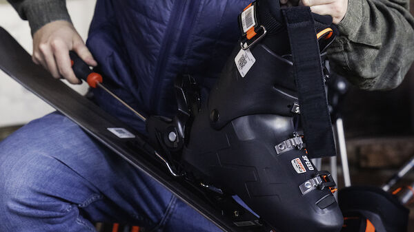 A person is adjusting their ski boot bindings on a ski, wearing a cap, blue vest, and jeans, and is kneeling in a ski equipment area.