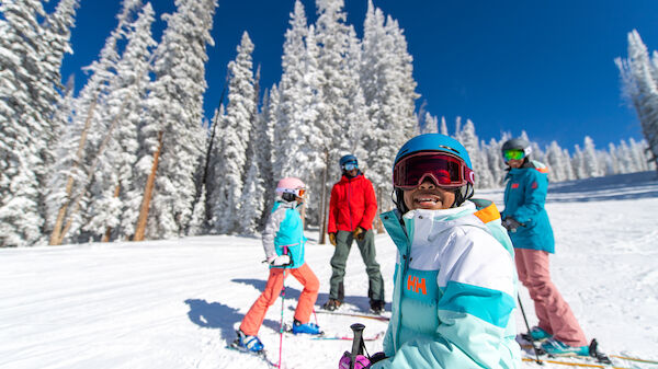 A group of people wearing colorful ski gear is standing and smiling on a snowy mountain slope, surrounded by snow-covered trees and clear blue sky.