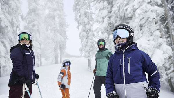 A group of people in winter clothing and ski gear stand on a snowy landscape surrounded by trees, looking ready to ski.