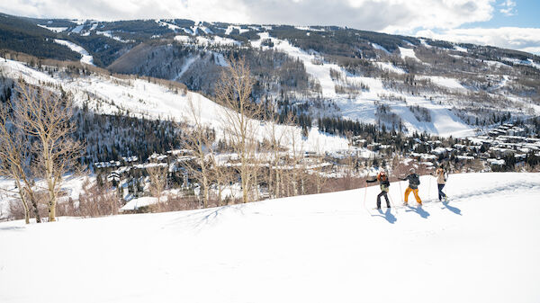 A snowy mountain landscape with three people skiing down the slope under a clear blue sky with scattered clouds, and trees in the foreground.