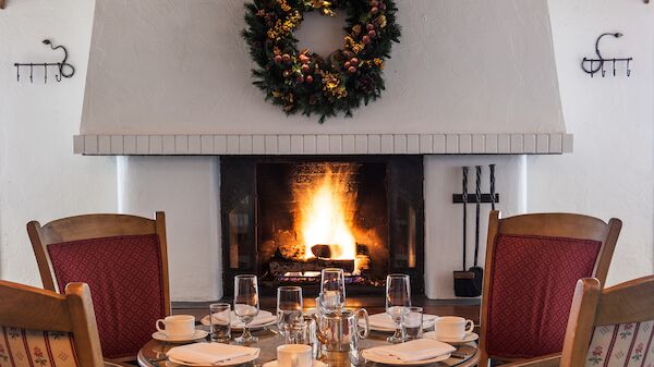 A cozy dining area with a festive wreath above a lit fireplace, surrounded by red chairs and a set table with glassware and dishes, creating a warm ambiance.