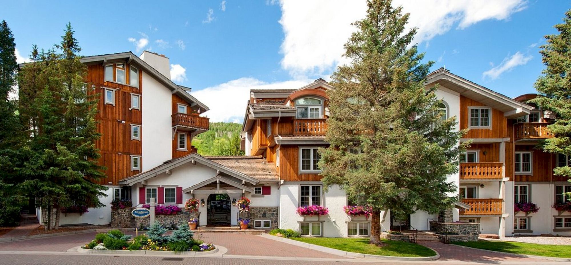 A charming lodge-style building with wooden accents, balconies, and greenery, set against a blue sky with fluffy clouds.