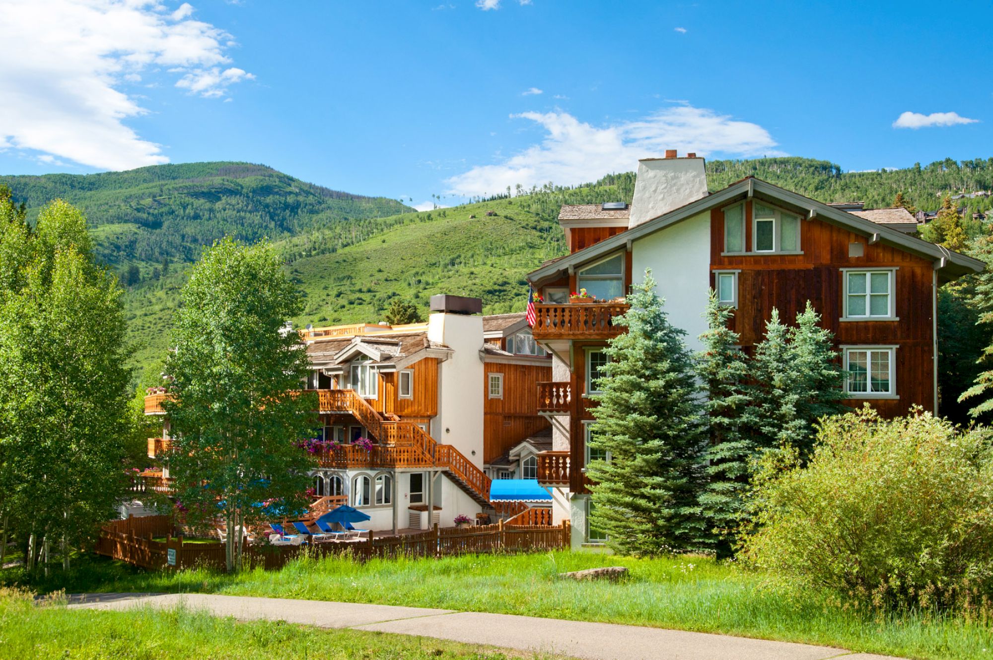 A picturesque mountain lodge surrounded by lush green trees and hills under a clear blue sky. The building features wooden balconies and chimneys.