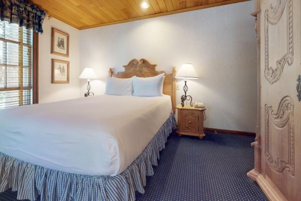 The image shows a cozy bedroom with a wooden ceiling, a double bed, two side tables with lamps, framed pictures on the wall, and a cupboard.