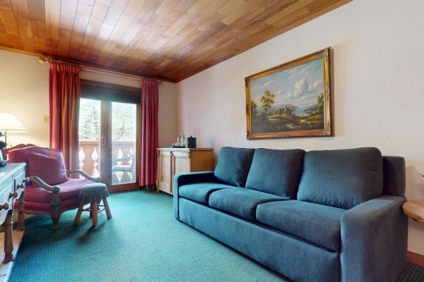 A cozy living room with a blue sofa, armchair, desk, lamp, and wall art. There is a window with red curtains and a view of greenery outside.