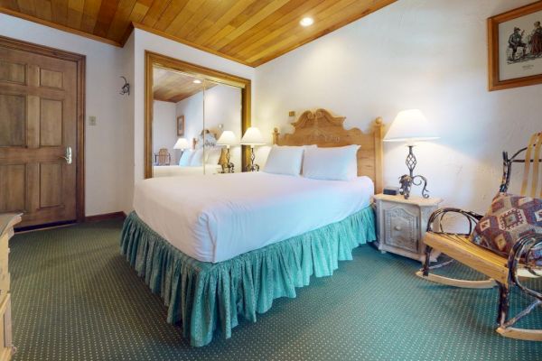 A cozy bedroom with a large bed, nightstands, lamps, a rocking chair, wooden ceiling, and a large mirror. There are paintings on the walls.