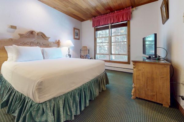 A cozy bedroom with a large bed, wooden furniture, a TV, a window with red curtains, and a green carpet. The walls are white.