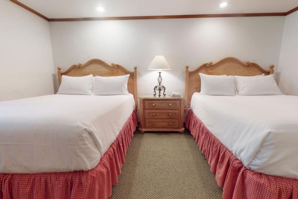 The image shows a bedroom with two neatly made beds, a shared nightstand with a lamp in the center, red bedskirts, and white walls.