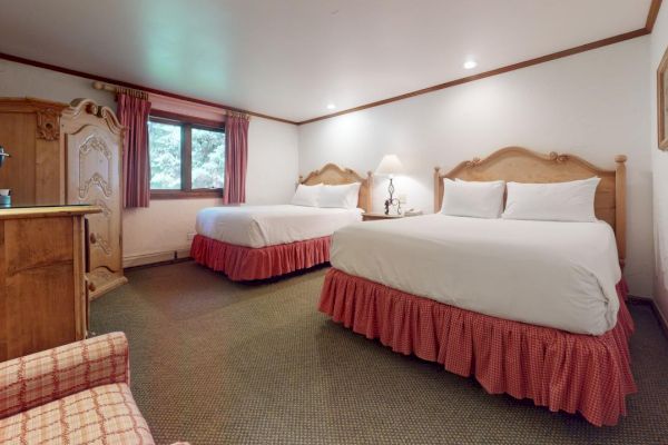 A cozy hotel room with two double beds, wooden furniture, a coffee maker, armchair, and a window with curtains letting in natural light.