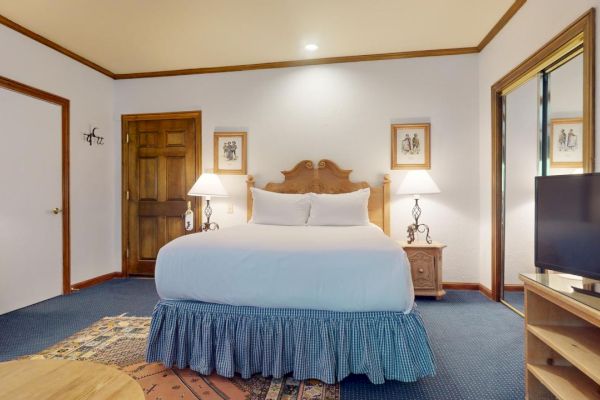 The image shows a well-lit bedroom with a large bed, bedside tables with lamps, a TV on a wooden shelf, and wall art.