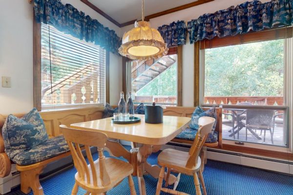 A cozy dining nook with a wooden table, chairs, and bench seat, a hanging light fixture, and windows with blue curtains showing an outdoor view.