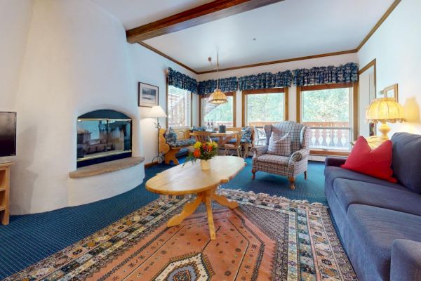 A cozy living room with a fireplace, TV, rug, couch, armchairs, a coffee table with flowers, a dining area, and a window with a view.