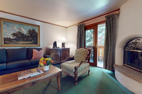 A cozy living room features a couch, armchair, coffee table with flowers, a painting, fireplace, sideboard, lamp, and glass doors with curtains.