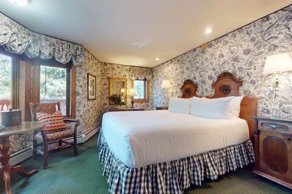 This image shows a cozy bedroom with a large bed, ornate wooden headboard, patterned wallpaper, two lamps, a seating area, and a window view.