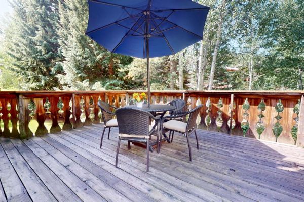 An outdoor wooden deck with a table, four chairs, and a blue umbrella; surrounded by trees and greenery.